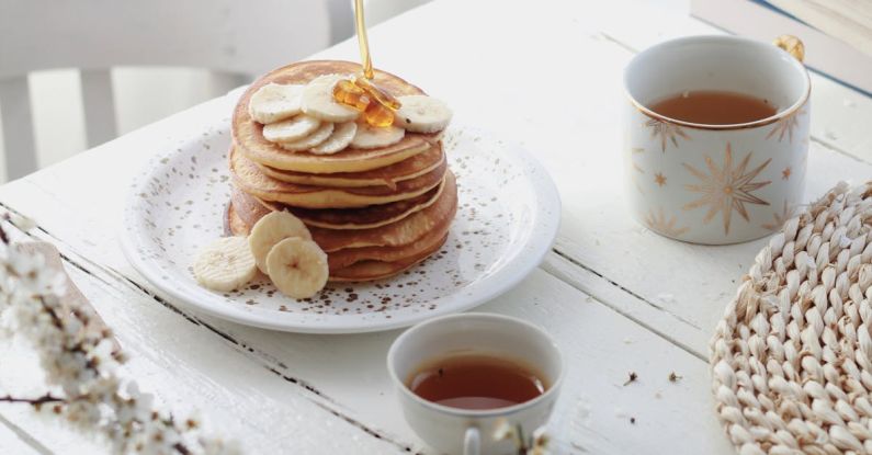 Energy-Efficient Kitchen - High angle of unrecognizable person adding caramel topping on tasty pancakes with banana slices served on table with cups of tea in morning