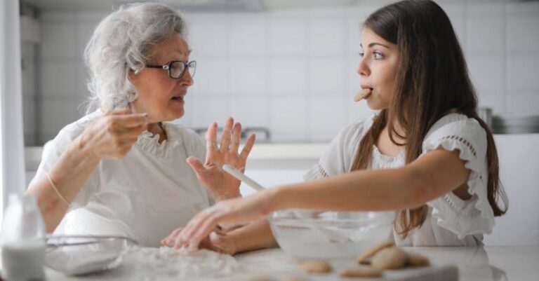 Eco-Friendly Kitchen - Calm senior woman and teenage girl in casual clothes looking at each other and talking while eating cookies and cooking pastry in contemporary kitchen at home